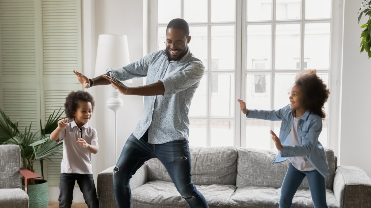 Happy African ethnicity father have fun teaches little preschool kids to dance in modern living room at home. Dad with son and daughter engaged in funny activity enjoy leisure carefree weekend concept