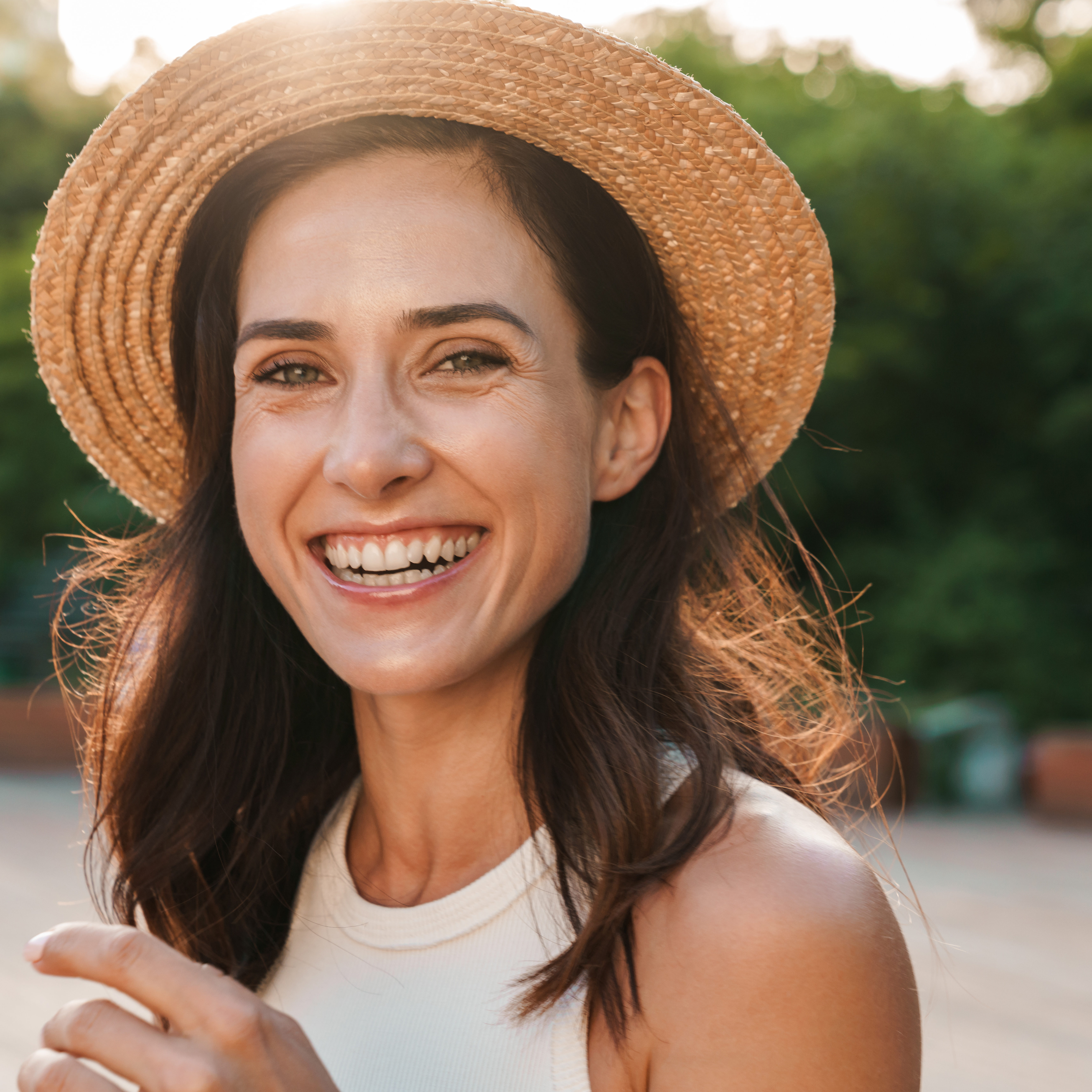 Image of beautiful middle-aged woman wearing straw hat laughing and looking at camera while walking in summer park